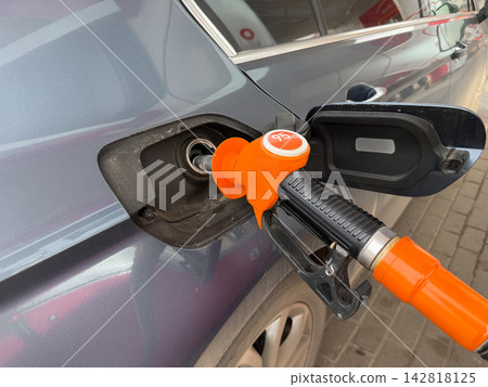 Fuel nozzle is positioned inside the gas tank of a blue vehicle at a gas station, with the fuel cap and pavement visible in the scene Fuel nozzle is positioned inside the gas tank of a blue vehicle at a gas station, with the fuel cap and pavement visible in the scene 142818125
