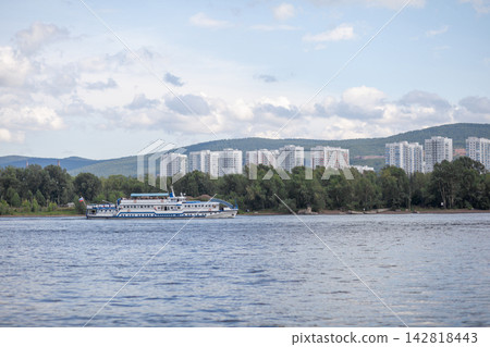 River cruise ship moves through tranquil waters with a backdrop of residential buildings and lush trees under a blue sky with clouds River cruise ship moves through tranquil waters with a backdrop of residential buildings and lush trees under a blue sky with clouds 142818443