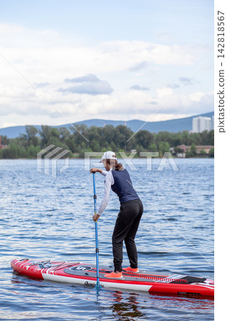 Woman paddleboards on a serene lake, dressed in athletic wear and a cap, with mountains and trees in the background Woman paddleboards on a serene lake, dressed in athletic wear and a cap, with mountains and trees in the background 142818567