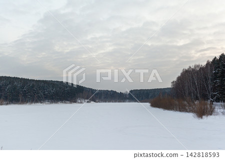 Snow-covered river landscape features a dense forest and cloudy sky, creating a serene winter scene with a frozen water surface 142818593