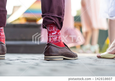 Individual in brown shoes and red patterned socks stands on cobblestone pavement, with blurred figures in the background at an outdoor gathering Individual in brown shoes and red patterned socks stands on cobblestone pavement, with blurred figures in the background at an outdoor gathering 142818607