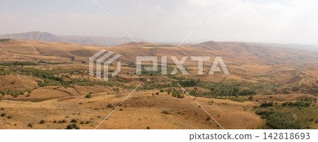 Expansive arid landscape features rolling hills and sparse vegetation with distant mountains under a cloudy sky in a rural area Expansive arid landscape features rolling hills and sparse vegetation with distant mountains under a cloudy sky in a rural area 142818693