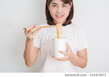 Happy young Asian woman enjoying a delicious cup of hot instant noodles while relaxing at home, Cheerful female eating instant ramen with a smile at apartment. Simple lifestyle and quick meal concept Happy young Asian woman enjoying a delicious cup of hot instant noodles while relaxing at home, Cheerful female eating instant ramen with a smile at apartment. Simple lifestyle and quick meal concept 142818971
