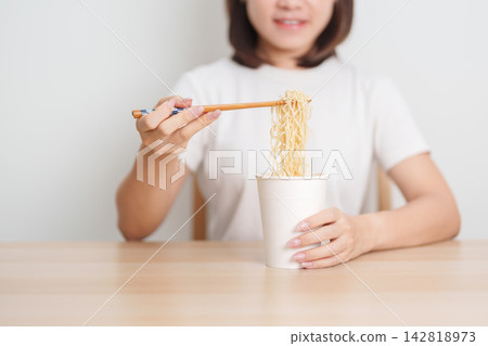 Happy young Asian woman enjoying a delicious cup of hot instant noodles while relaxing at home, Cheerful female eating instant ramen with a smile at apartment. Simple lifestyle and quick meal concept 142818973