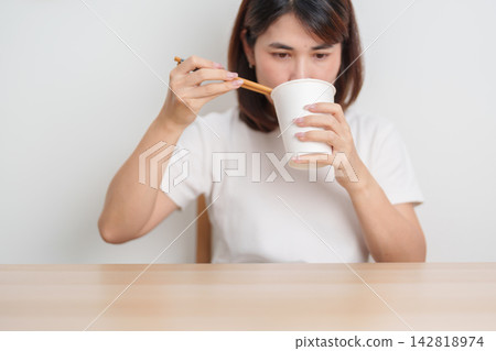 Happy young Asian woman enjoying a delicious cup of hot instant noodles while relaxing at home, Cheerful female eating instant ramen with a smile at apartment. Simple lifestyle and quick meal concept Happy young Asian woman enjoying a delicious cup of hot instant noodles while relaxing at home, Cheerful female eating instant ramen with a smile at apartment. Simple lifestyle and quick meal concept 142818974