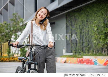 A smiling businesswoman helmeted and holding her smartphone enjoys a bike ride. Her expression conveys the modern concept of work-life balance and the freedom to stay connected. A smiling businesswoman helmeted and holding her smartphone enjoys a bike ride. Her expression conveys the modern concept of work-life balance and the freedom to stay connected. 142819152