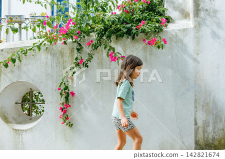 Small Girl Walking Along a Wall with Circular Green Window and Pink Bougainvillea Small Girl Walking Along a Wall with Circular Green Window and Pink Bougainvillea 142821674