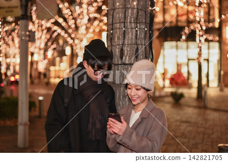 一對年輕夫婦在掛滿聖誕彩燈的街道上低頭看著他們的智慧型手機。 142821705