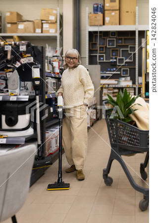Senior woman smiling, holding a vacuum cleaner while shopping in a retail store Senior woman smiling, holding a vacuum cleaner while shopping in a retail store 142824046