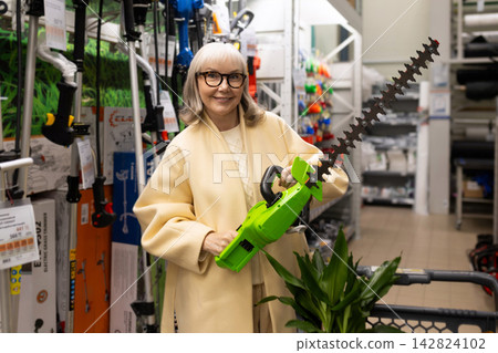 Senior woman smiling, holding a hedge trimmer and shopping in a hardware store 142824102