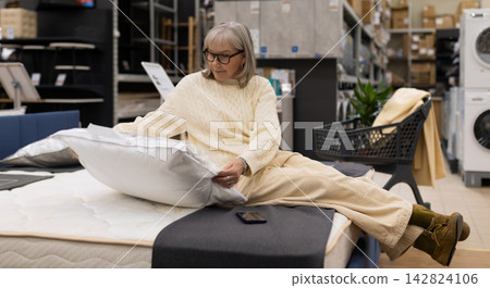 Mature woman testing a pillow on a mattress while shopping for home goods Mature woman testing a pillow on a mattress while shopping for home goods 142824106