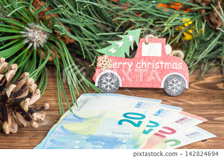 Euro banknotes on a wooden table with a pine cone, pine branch, and a red Christmas truck ornament. 142824694