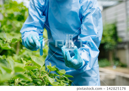 Agronomist in a protective suit holding a test tube and flask in a green garden. Outdoor laboratory research focusing 142825176