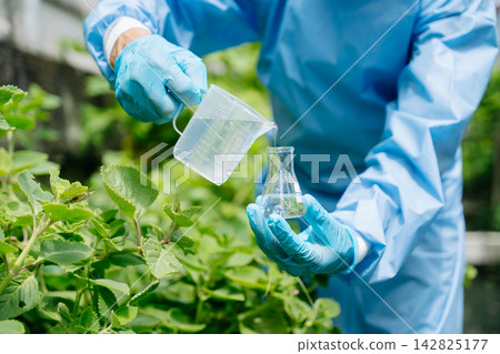Agronomist in a protective suit holding a test tube and flask in a green garden. Outdoor laboratory research focusing Agronomist in a protective suit holding a test tube and flask in a green garden. Outdoor laboratory research focusing 142825177