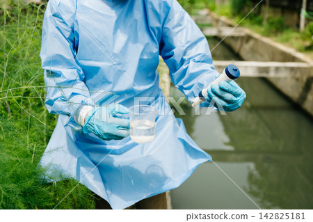 Environmental researcher in blue gown testing water purity in a beaker using a digital meter 142825181