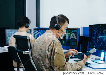 Senior military officer in camouflage using a tablet to analyze tactical radar data alongside his team in a modern cybersecurity command center. Senior military officer in camouflage using a tablet to analyze tactical radar data alongside his team in a modern cybersecurity command center. 142825267