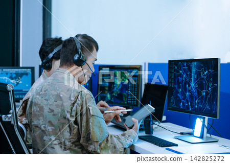 Senior military officer in camouflage using a tablet to analyze tactical radar data alongside his team in a modern cybersecurity command center. Senior military officer in camouflage using a tablet to analyze tactical radar data alongside his team in a modern cybersecurity command center. 142825272