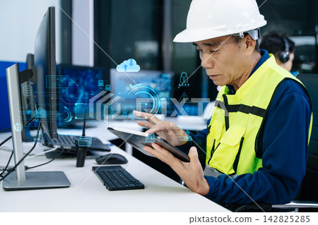 Asian engineer in a safety vest and headset analyzing clean energy HUDs and robotic automation interfaces in high tech industrial control room. 142825285