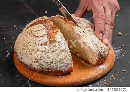 Freshly baked round bread loaf being sliced with a knife on a wooden cutting board, flour dust visible on the surface and around the loaf Freshly baked round bread loaf being sliced with a knife on a wooden cutting board, flour dust visible on the surface and around the loaf 142825971