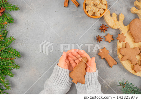 Hands holding a gingerbread Christmas tree cookie surrounded by festive decorations, including star anise, sugar cubes, and pine branches on a gray background Hands holding a gingerbread Christmas tree cookie surrounded by festive decorations, including star anise, sugar cubes, and pine branches on a gray background 142825979