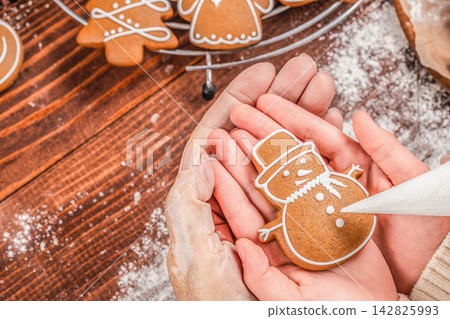 Hands of a child and adult decorating a gingerbread snowman cookie with icing on a wooden table, surrounded by various shaped cookies and flour 142825993