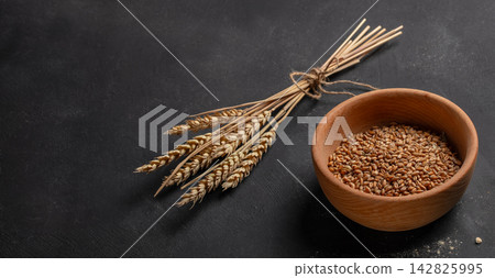 Wheat grains in a wooden bowl beside a bundle of wheat stalks on a dark textured surface, showcasing natural agricultural elements and rustic kitchen decor 142825995