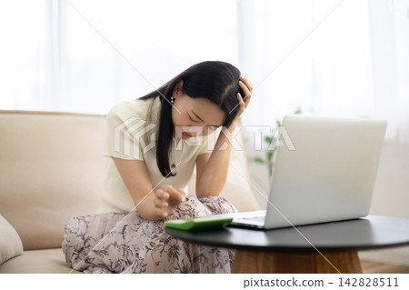 A young Japanese woman is sitting on a sofa by the window, using a laptop and calculator, holding her head in her hands. A young Japanese woman is sitting on a sofa by the window, using a laptop and calculator, holding her head in her hands. 142828511