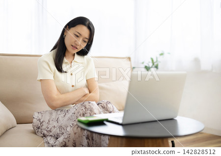 A young Japanese woman sits on a sofa by the window, arms crossed, tilting her head while looking at her laptop. A young Japanese woman sits on a sofa by the window, arms crossed, tilting her head while looking at her laptop. 142828512