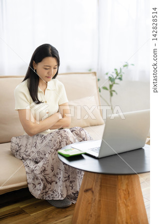 A young Japanese woman sits on a sofa by the window, arms crossed, tilting her head while looking at her laptop. A young Japanese woman sits on a sofa by the window, arms crossed, tilting her head while looking at her laptop. 142828514