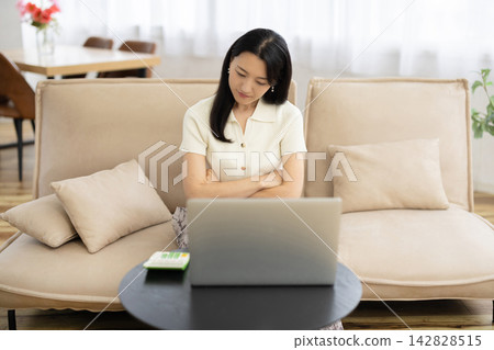 A young Japanese woman sits on a sofa by the window, arms crossed, tilting her head while looking at her laptop. A young Japanese woman sits on a sofa by the window, arms crossed, tilting her head while looking at her laptop. 142828515