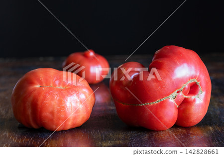 Three unusual, naturally misshapen red tomatoes on rustic wooden table. Tomatoes have pronounced irregular shapes, deep grooves, scars, surface cracking. Food that does not meet standard expectations Three unusual, naturally misshapen red tomatoes on rustic wooden table. Tomatoes have pronounced irregular shapes, deep grooves, scars, surface cracking. Food that does not meet standard expectations 142828661