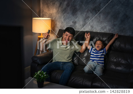 cheerful father and daughter watching sport tv and celebration with arm raised in the living room at night cheerful father and daughter watching sport tv and celebration with arm raised in the living room at night 142828819
