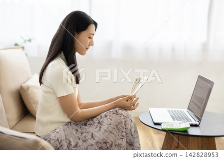 一位年輕的日本女子坐在窗邊的沙發上,一邊看著筆記型電腦,一邊數著一疊疊的日圓鈔票。 一位年輕的日本女子坐在窗邊的沙發上,一邊看著筆記型電腦,一邊數著一疊疊的日圓鈔票。 142828953