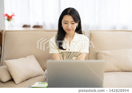 A young Japanese woman counts stacks of Japanese yen bills while looking at her laptop on a sofa by the window. A young Japanese woman counts stacks of Japanese yen bills while looking at her laptop on a sofa by the window. 142828954