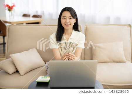 A young Japanese woman smiles at the camera, looking at a laptop and a wad of Japanese yen bills while sitting on a sofa by the window. A young Japanese woman smiles at the camera, looking at a laptop and a wad of Japanese yen bills while sitting on a sofa by the window. 142828955