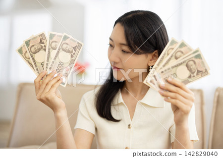 一位年輕的日本女子坐在沙發上，面帶微笑，手裡拿著一沓日元鈔票。 142829040