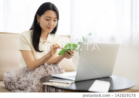 一位年輕的日本女性在桌子前使用計算器，桌子旁放著筆記型電腦、存摺和一張10,000日圓的鈔票。 142829178