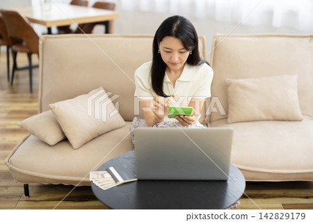 一位年輕的日本女性在桌子前使用計算器,桌子旁放著筆記型電腦、存摺和一張10,000日圓的鈔票。 一位年輕的日本女性在桌子前使用計算器,桌子旁放著筆記型電腦、存摺和一張10,000日圓的鈔票。 142829179