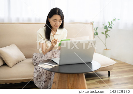 一位年輕的日本女性在桌子前使用計算器,桌子旁放著筆記型電腦、存摺和一張10,000日圓的鈔票。 一位年輕的日本女性在桌子前使用計算器,桌子旁放著筆記型電腦、存摺和一張10,000日圓的鈔票。 142829180