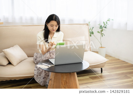 一位年輕的日本女性在桌子前使用計算器,桌子旁放著筆記型電腦、存摺和一張10,000日圓的鈔票。 一位年輕的日本女性在桌子前使用計算器,桌子旁放著筆記型電腦、存摺和一張10,000日圓的鈔票。 142829181