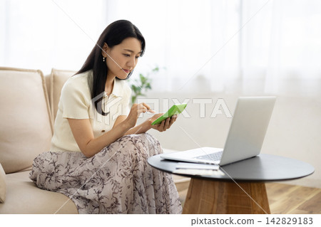 一位年輕的日本女性在桌子前使用計算器，桌子旁放著筆記型電腦、存摺和一張10,000日圓的鈔票。 142829183