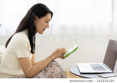 一位年輕的日本女性在桌子前使用計算器，桌子旁放著筆記型電腦、存摺和一張10,000日圓的鈔票。 142829184