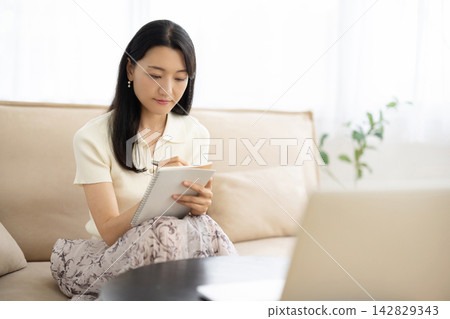 A young Japanese woman is sitting on a sofa by the window, taking notes on her laptop. A young Japanese woman is sitting on a sofa by the window, taking notes on her laptop. 142829343