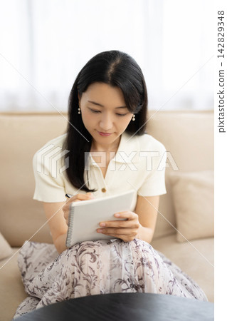 一位年輕的日本女子坐在窗邊的沙發上，用筆記型電腦做筆記。 142829348