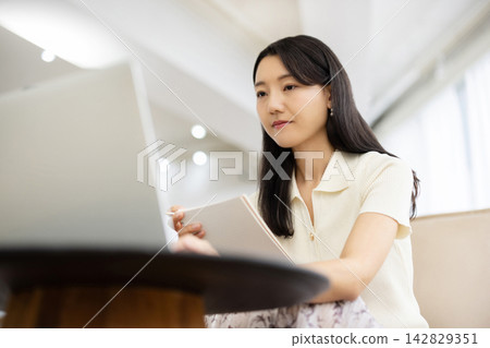 A young Japanese woman is sitting on a sofa by the window, taking notes on her laptop. 142829351