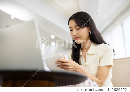 A young Japanese woman is sitting on a sofa by the window, taking notes on her laptop. A young Japanese woman is sitting on a sofa by the window, taking notes on her laptop. 142829352