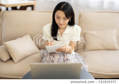A young Japanese woman is sitting on a sofa by the window, taking notes on her laptop. A young Japanese woman is sitting on a sofa by the window, taking notes on her laptop. 142829355