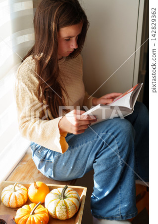 Teen girl reading book by window with pumpkins in autumn light Teen girl reading book by window with pumpkins in autumn light 142829362