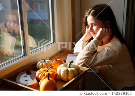 Teen girl relaxing by window with decorative pumpkins at home Teen girl relaxing by window with decorative pumpkins at home 142829363