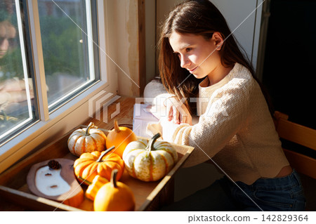 Teen girl relaxing by window with decorative pumpkins at home Teen girl relaxing by window with decorative pumpkins at home 142829364